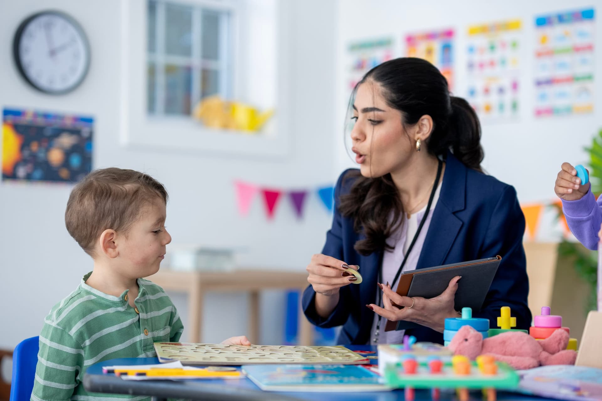 BCBA clinician in professional attire conducting a developmental skills assessment with a young toddler boy at a table, using a letter puzzle board and holding a tablet in a bright educational therapy room