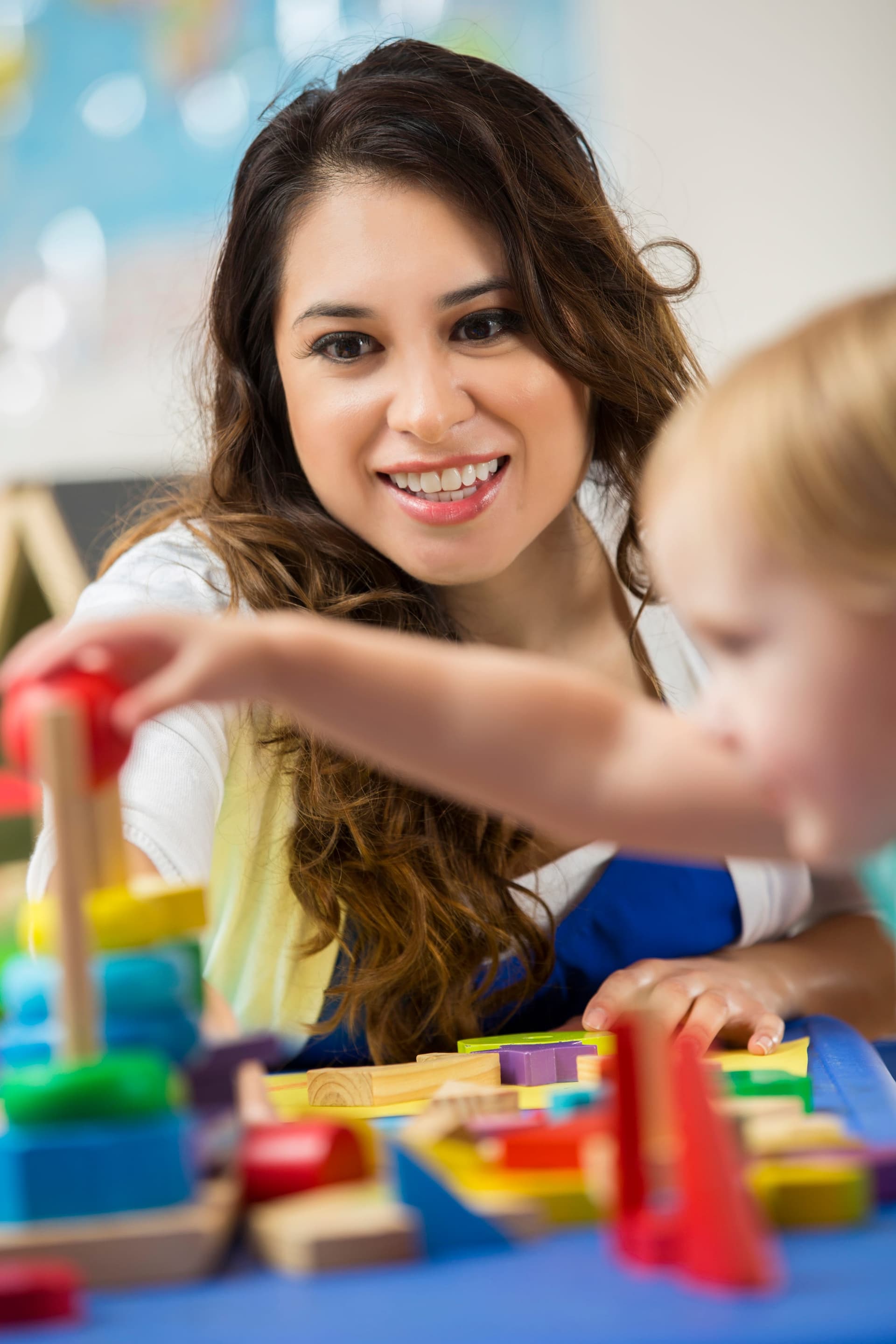 Latina behavior therapist smiling and making eye contact while reaching toward colorful stacking blocks with a young toddler during a naturalistic play-based ABA session in a bright educational setting