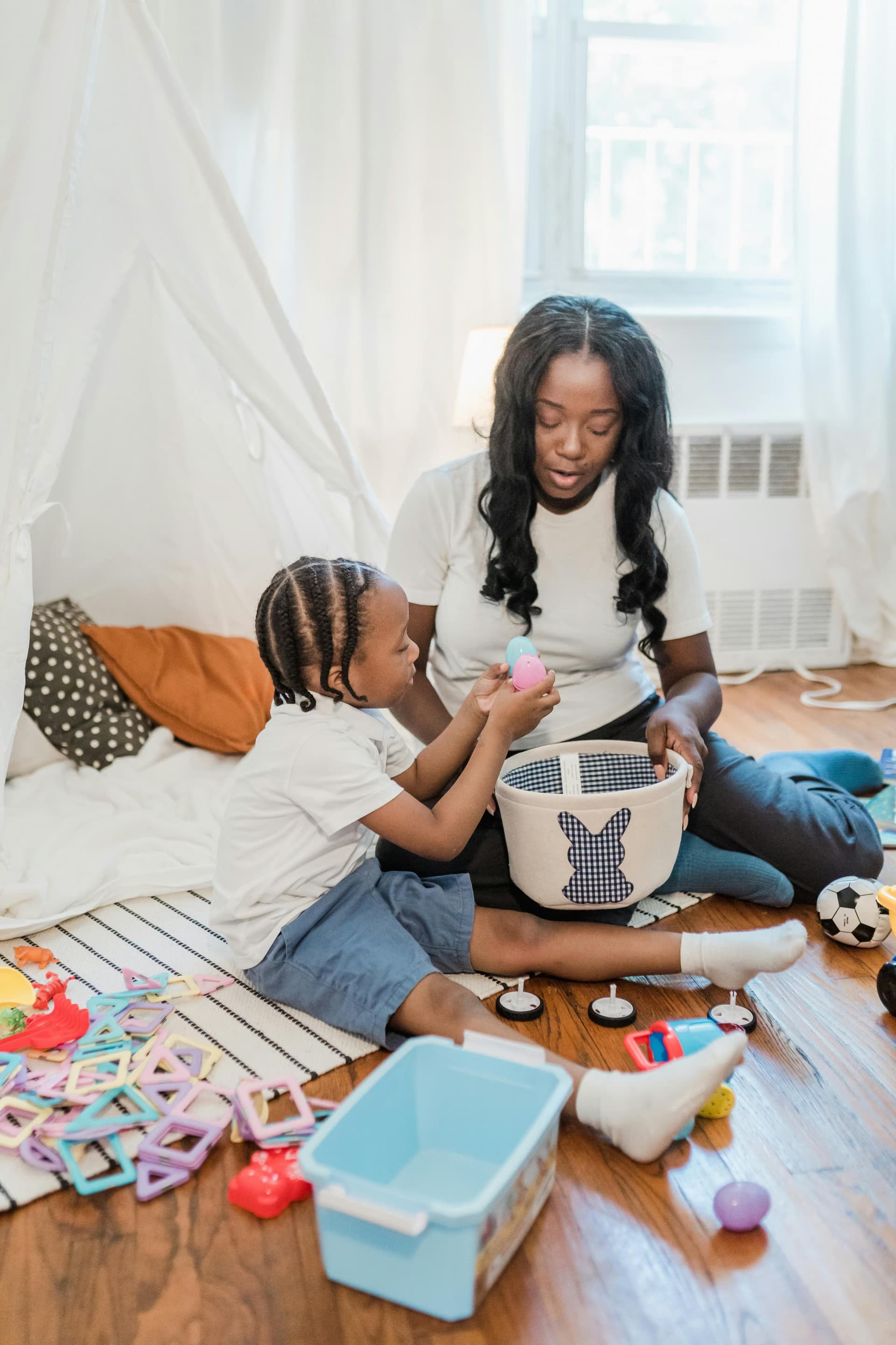 Black mother sitting on the floor with her young son engaged in naturalistic play, handing him a colorful egg from a basket surrounded by toys in a warm home environment