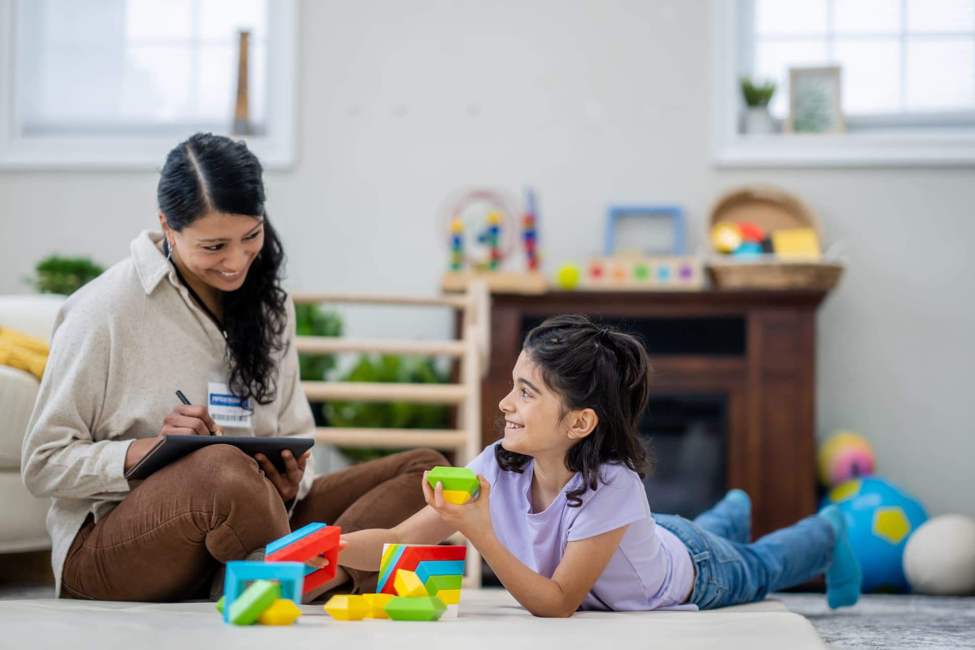 Behavior technician sitting on the floor with a young Hispanic girl, using colorful building blocks for skill acquisition while recording data on a tablet