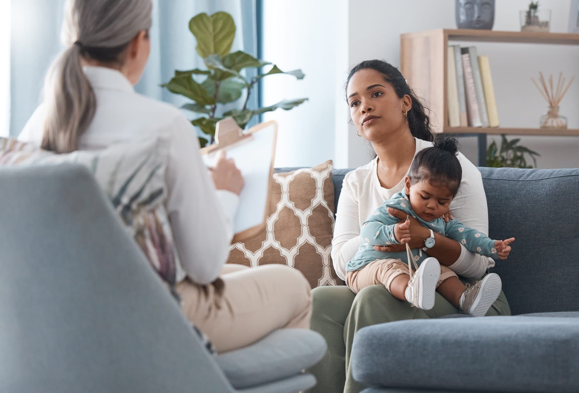 Latina mother holding her infant during an intake consultation with a clinician taking notes on a clipboard in a home setting
