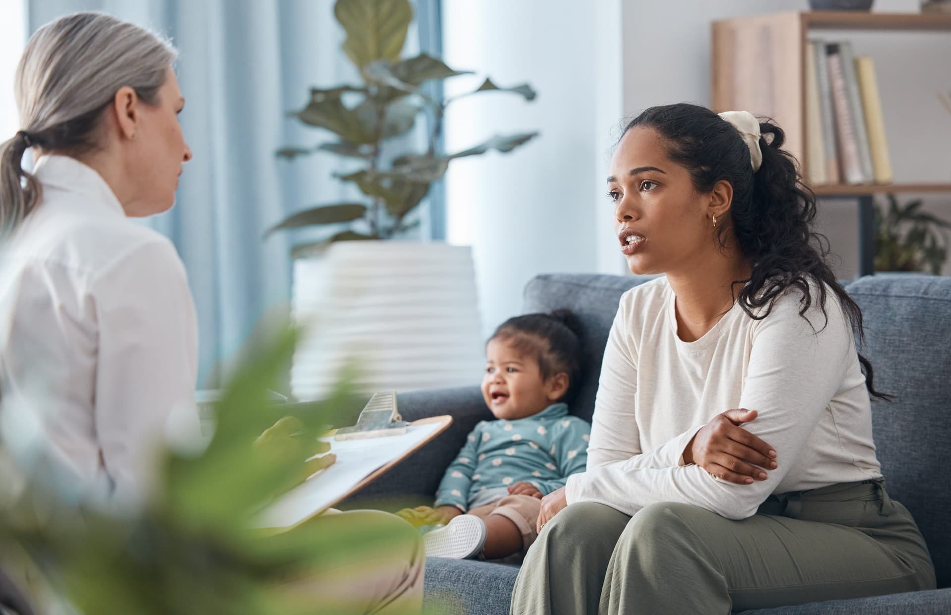 BCBA clinician consulting with a mother and her infant on a home visit, reviewing documentation on a clipboard while the baby smiles between them