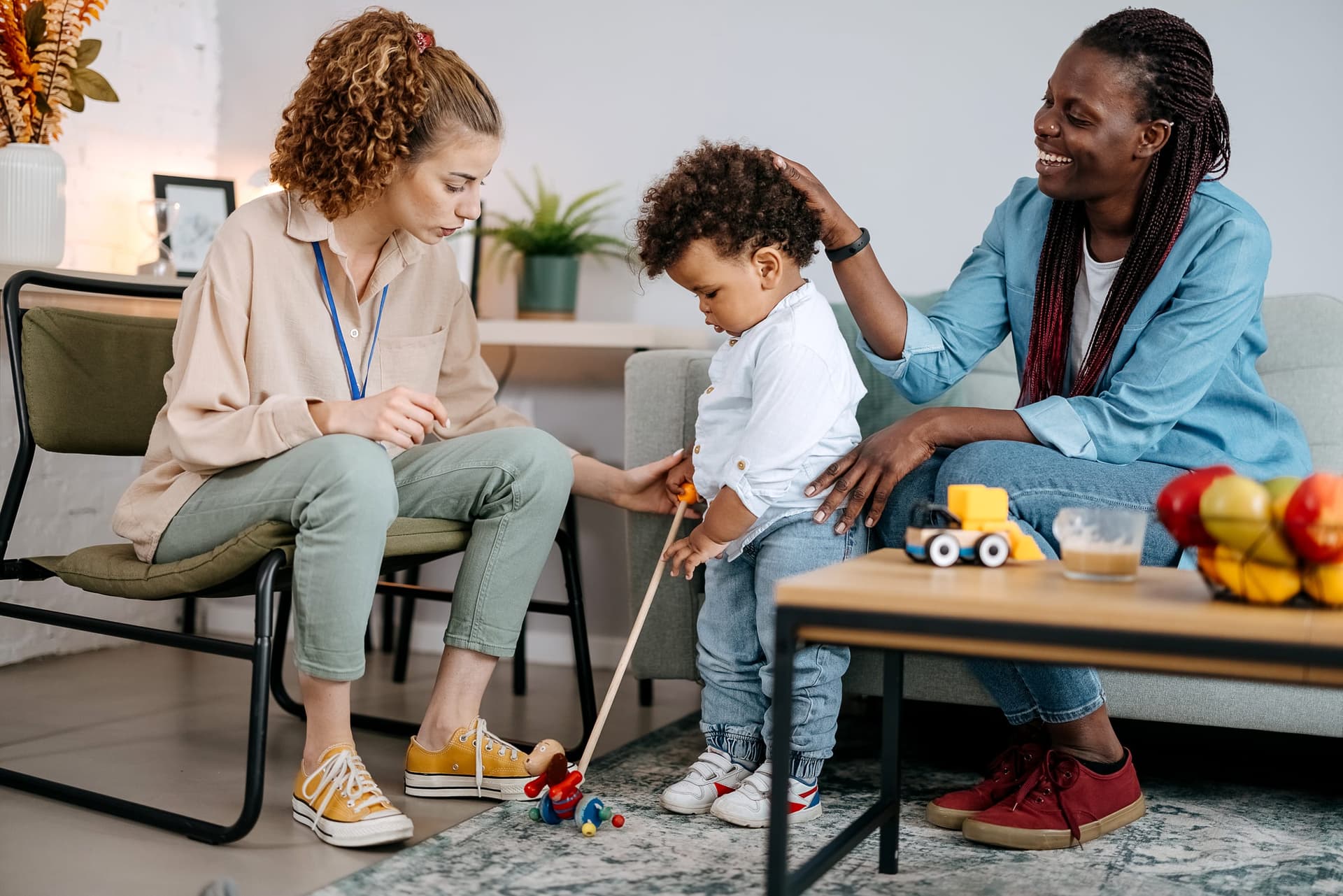 ABA clinician with a visible badge guiding a toddler with a wooden push toy during a naturalistic teaching activity while the child's mother watches and smiles in a home-based parent training session