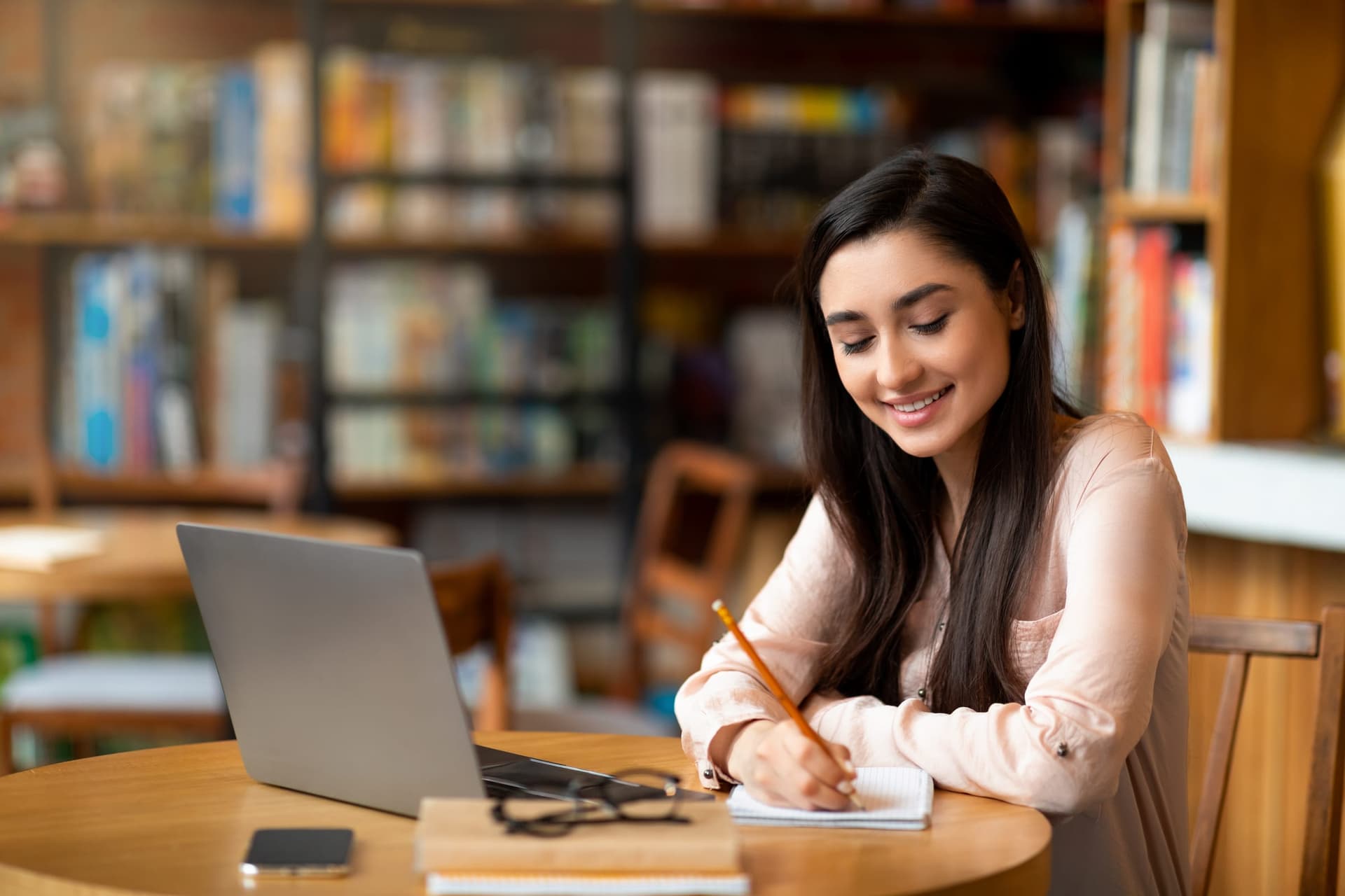 A student sitting at a round wooden table in a library, writing in a spiral notebook with a pencil beside an open laptop, with reading glasses and a phone on the table and shelves of books behind her