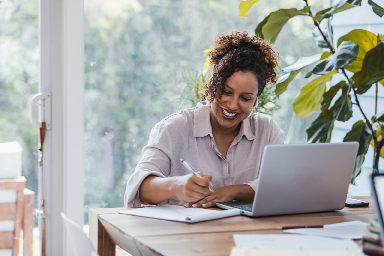 Smiling woman with curly hair writing notes beside an open laptop at a wooden desk in a bright, plant-filled home workspace